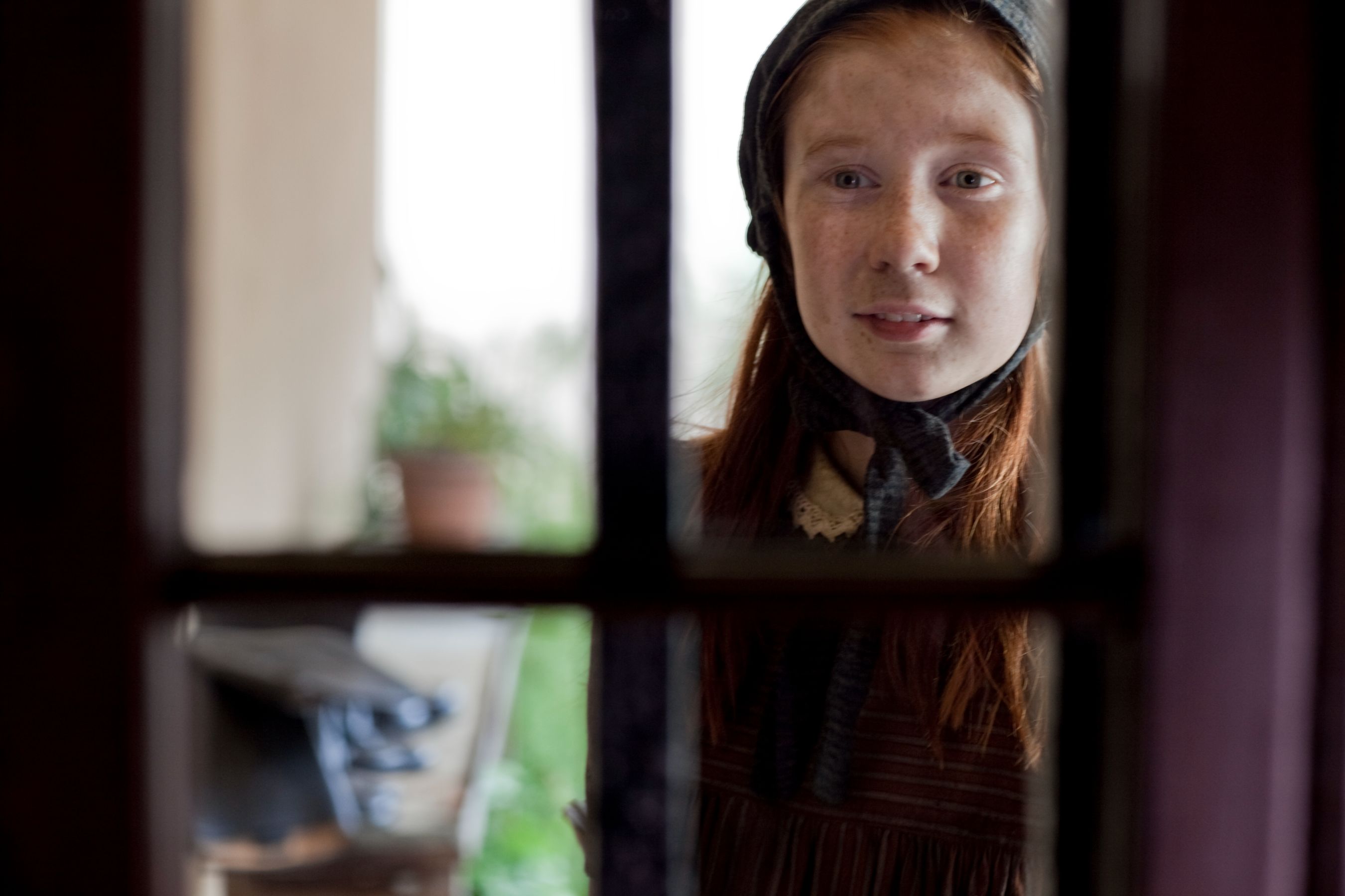 A young girl with long reddish hair, wearing a brown dress and dark cape, stands outside a window, looking in with a gentle smile. The scene is softly illuminated, with green plants visible in the background.