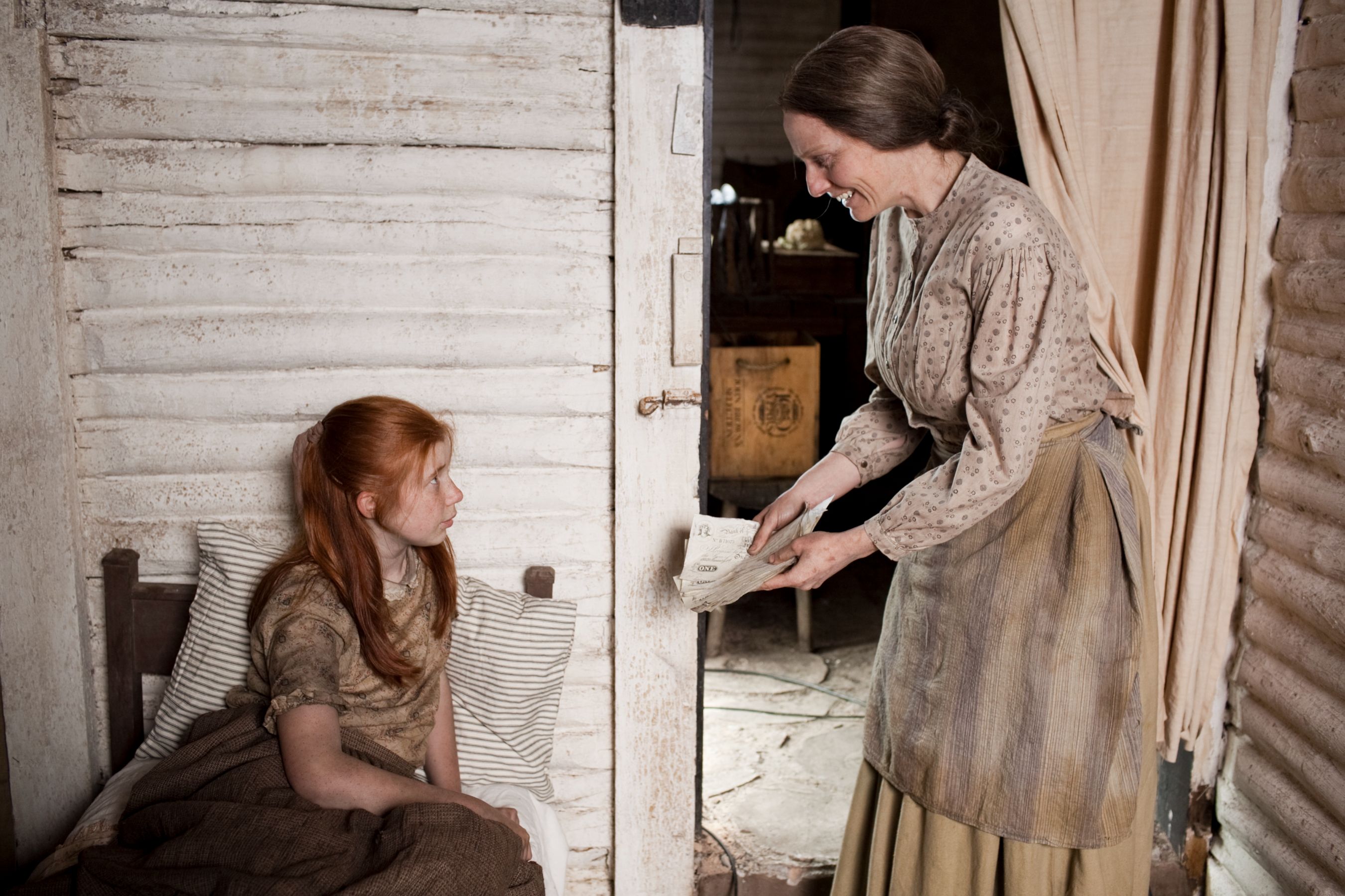 A woman in period attire smiles as she hands a letter to a young girl with red hair, who sits on a bed looking curiously at her. The interior is rustic, with wooden walls and simple furnishings, conveying a historical setting.