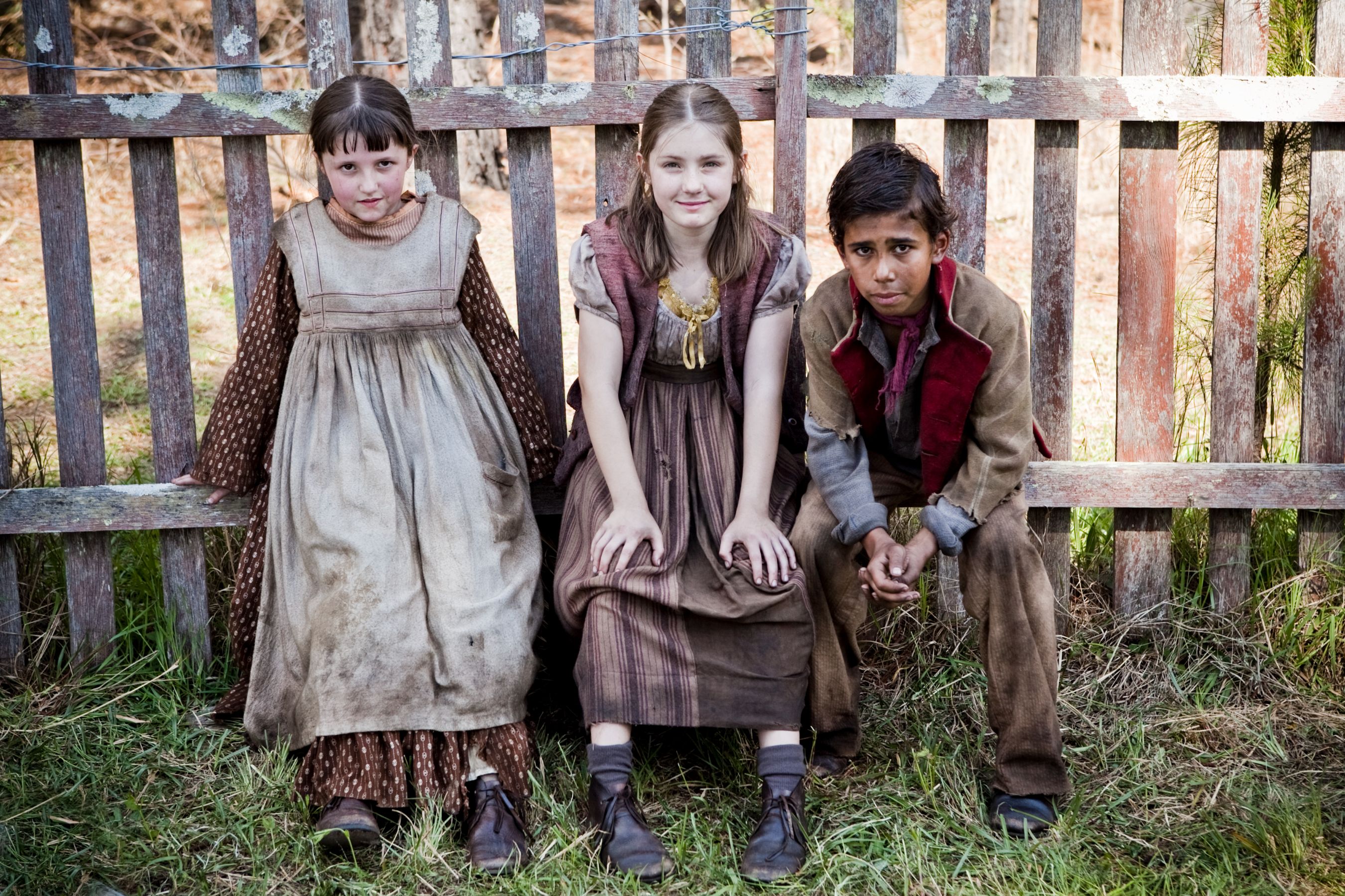 Three children are seated near a wooden fence, dressed in vintage, worn clothing. The two girls on the left wear long, modest dresses, while the boy on the right sits in a more rugged outfit, all appearing to be from a past era. They share a casual pose, reflecting a sense of camaraderie amidst a rustic outdoor setting.