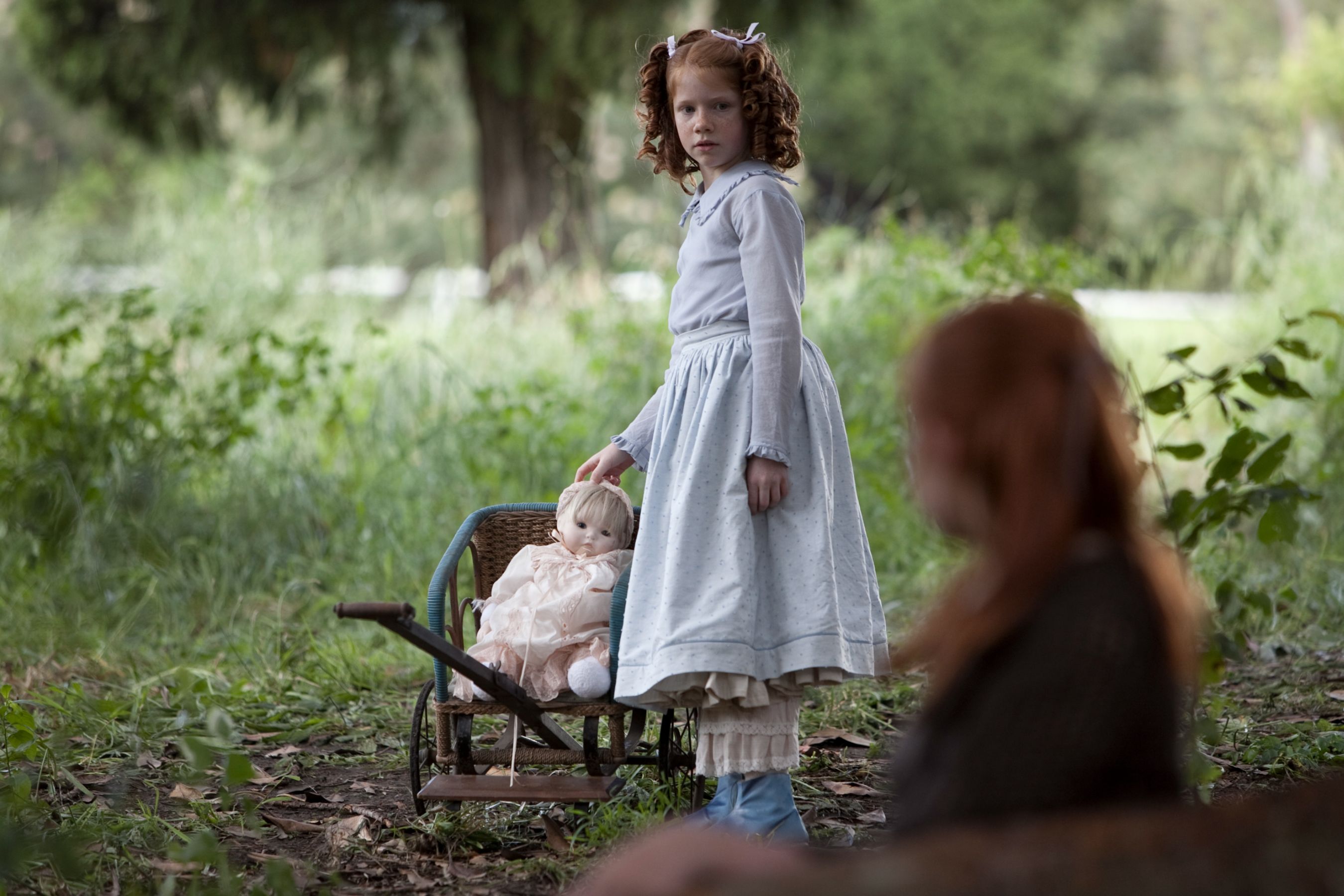 A young girl with curly hair and a blue dress stands beside a small doll carriage, gently touching the doll inside. In the background, another girl with reddish hair is partially visible, seated on the ground among lush greenery.