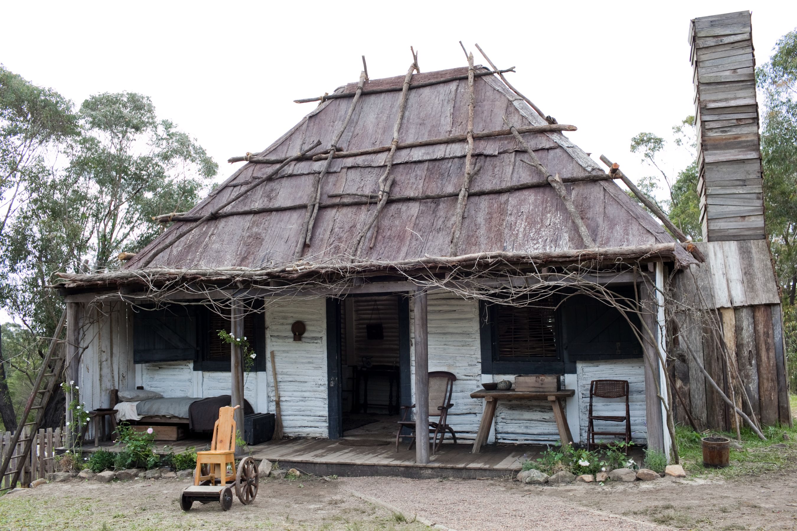 A rustic wooden cottage features a thatched roof made from branches and straw. The front porch has a small table and chairs, with a cart in the foreground and surrounding greenery. A tall, narrow chimney rises from the side of the house, blending into a serene natural setting.