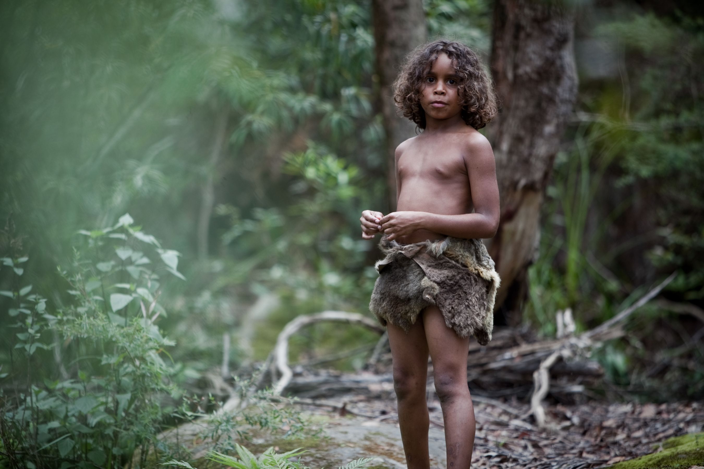 A young child stands in a lush, green forest, wearing a simple garment made from animal hide. The child has curly hair and appears to be observing the surroundings with a curious expression.