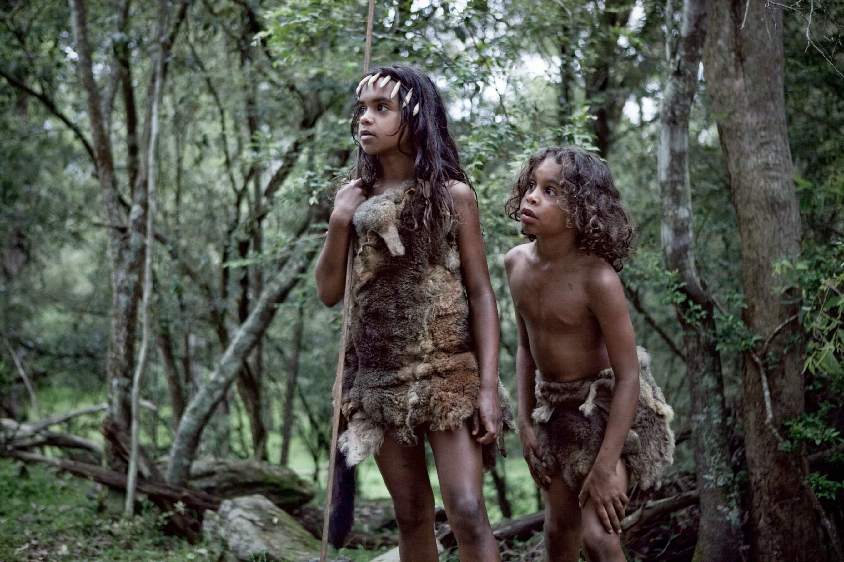 Two young children are dressed in primitive clothing made from animal skins, standing in a dense forest surrounded by trees. One child holds a spear and looks attentively at something in the distance, while the other appears curious, observing their surroundings.