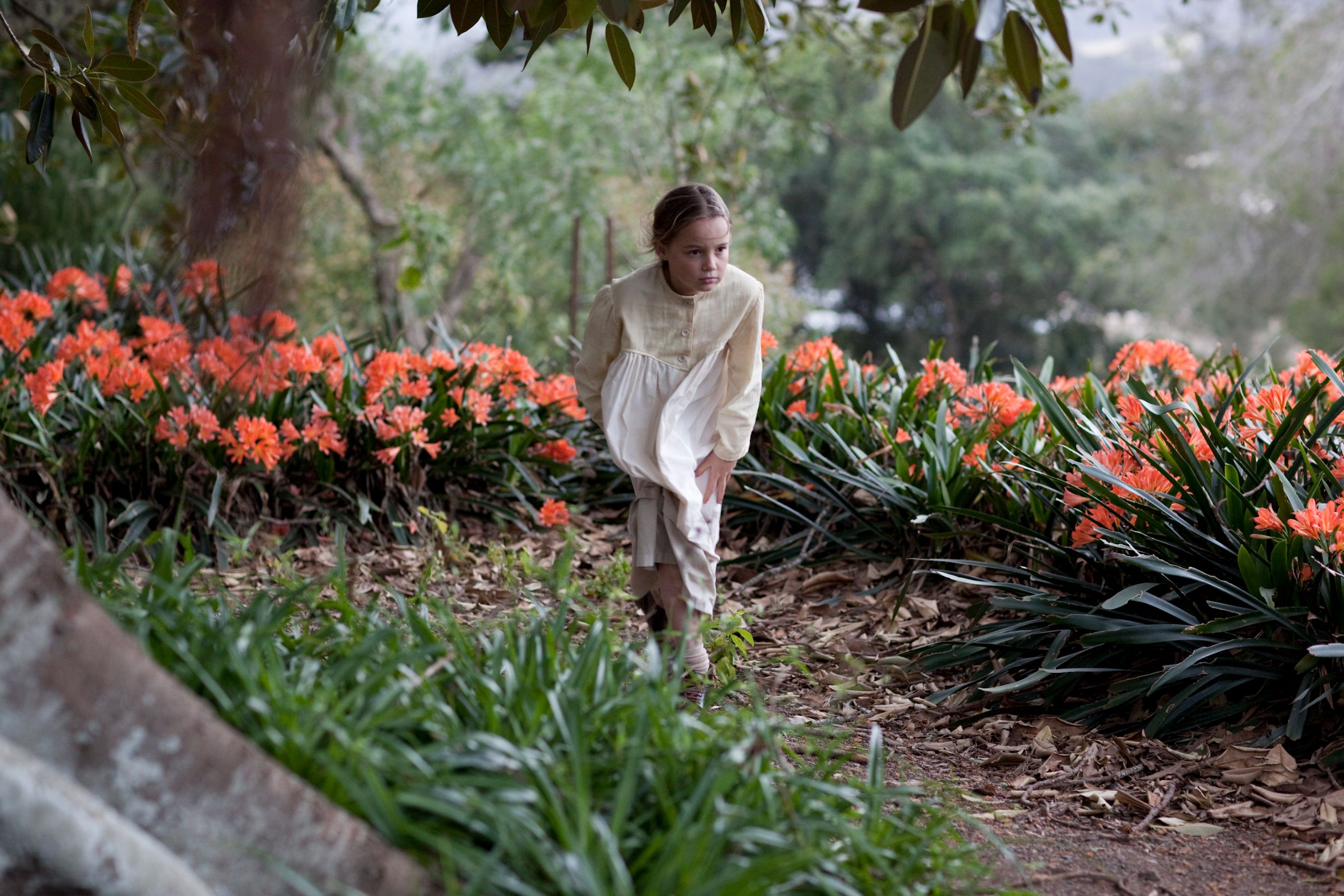 A young girl wearing a light-colored dress kneels among vibrant orange flowers in a garden. She appears focused, surrounded by lush greenery and softly blurred trees in the background.