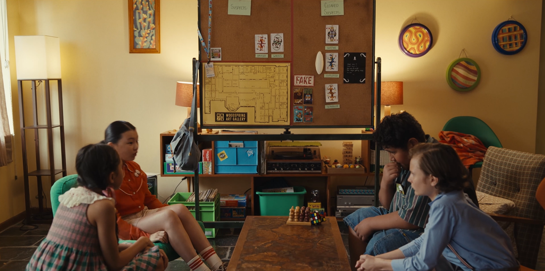 A group of four children is seated in a cozy room, discussing strategies around a wooden table. A large bulletin board displays a map and a collection of notes and cards, while colorful storage bins and a bookshelf can be seen in the background. The atmosphere is casual and focused, suggesting they are engaged in a game or problem-solving activity.
