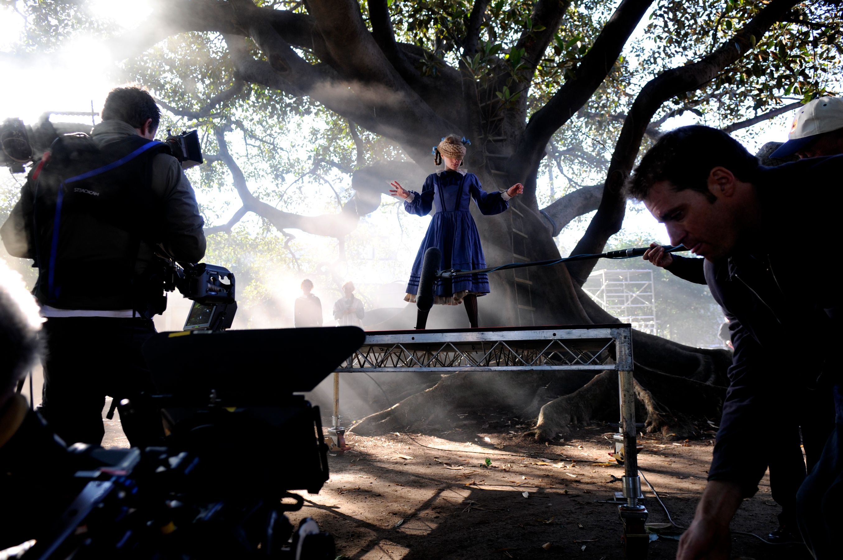 A child in a blue dress stands on a platform, performing theatrically under a large tree. Surrounding her, crew members operate cameras and equipment, with mist swirling in the background, suggesting an outdoor film or theater production.