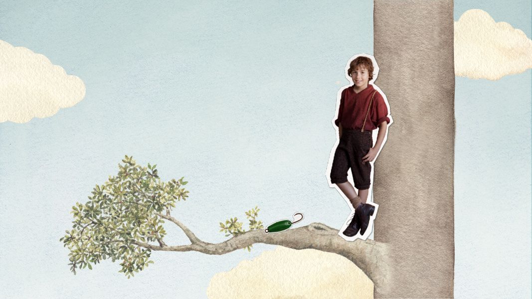 A young boy with curly red hair wears a red shirt and black shorts, standing on a tree branch. The background features a light blue sky with fluffy white clouds, and a green object is visible on the branch beside him.