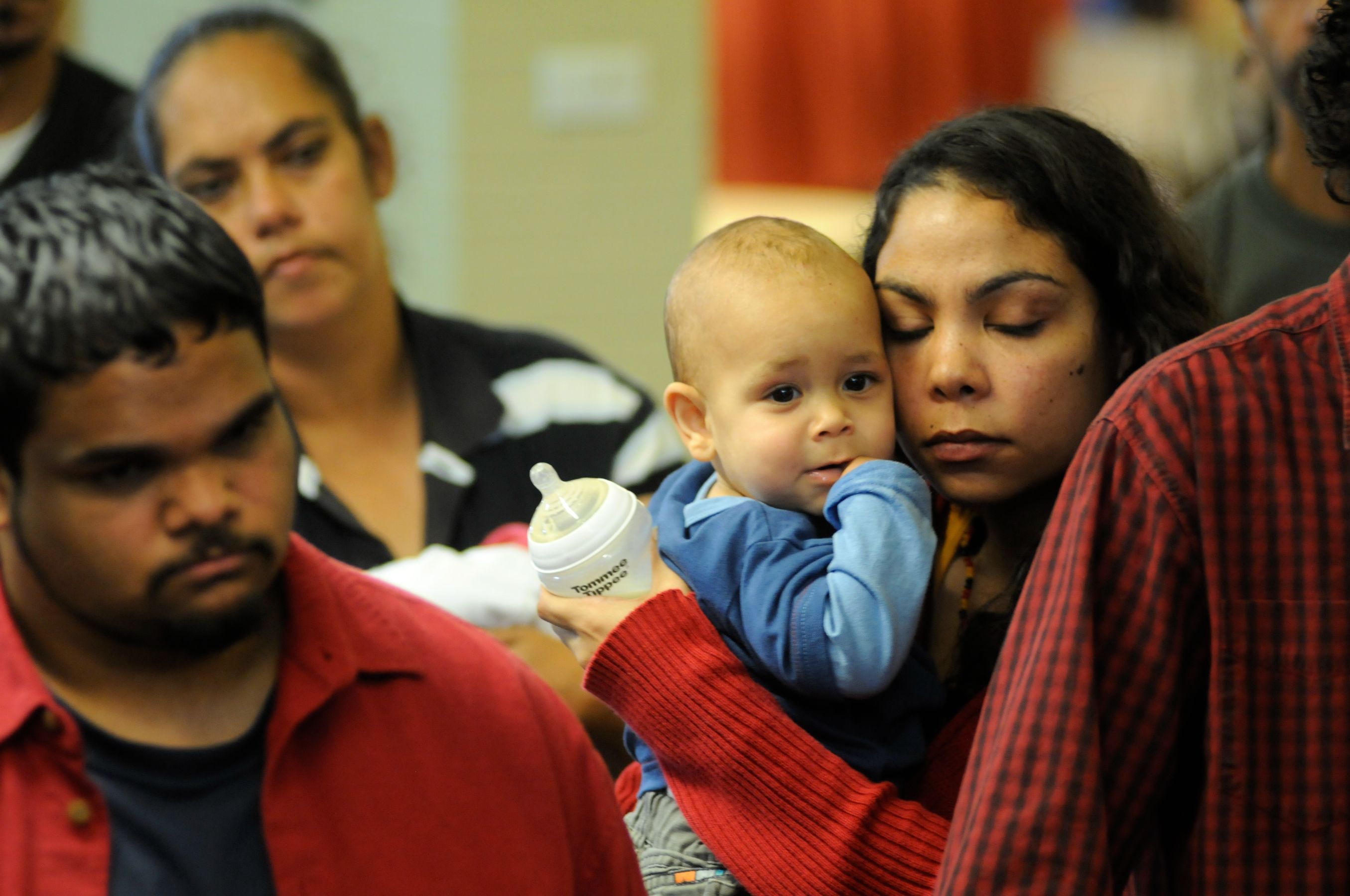 A woman holds a small child closely while closing her eyes in a contemplative expression, conveying warmth and care. In the background, another individual looks on with a serious demeanor, and other people are partially visible, adding to the sense of the moment.