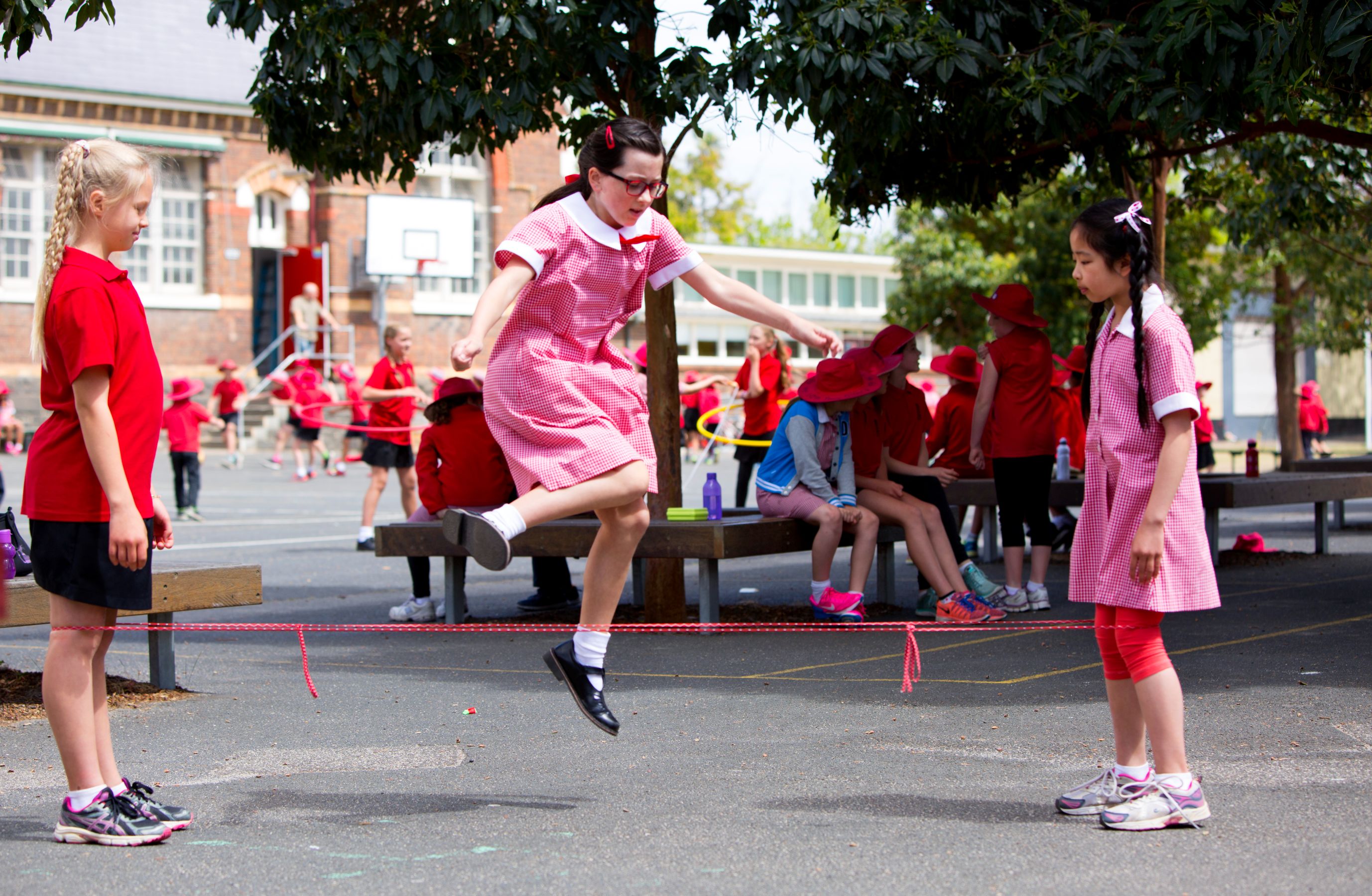 A girl in a pink dress and glasses jumps rope while another girl in a red shirt stands by, watching. In the background, other children in red clothing are engaged in various activities, and benches are visible under leafy trees.