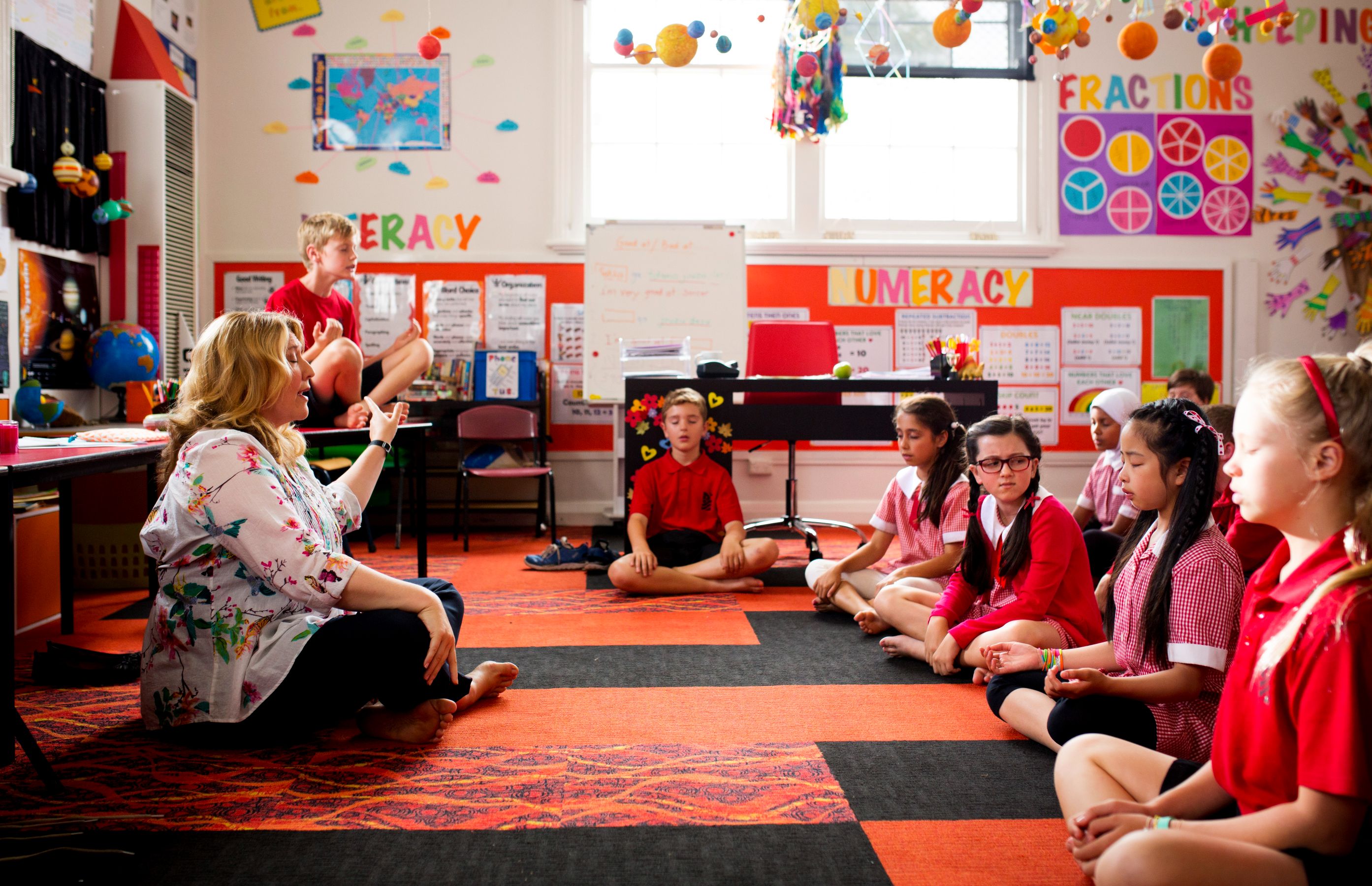 A group of children sits cross-legged on a colorful carpet in a classroom, engaged in meditation. A teacher, seated nearby and gesturing, guides them as a boy balances on a chair in the background. Brightly colored educational posters adorn the walls, and various classroom supplies are visible on tables.