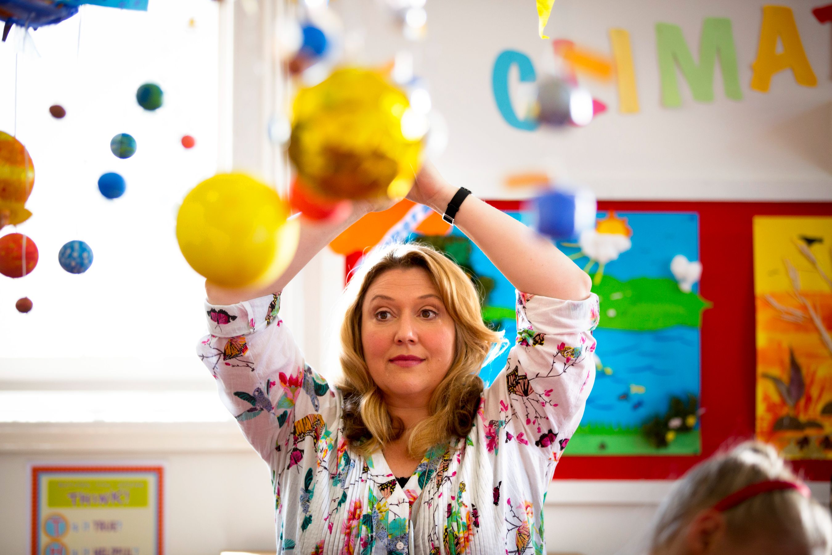A woman with blonde, wavy hair is in a colorful classroom filled with hanging decorations of planets and celestial bodies. She is reaching upwards with a focused expression, while vibrant artwork adorns the walls behind her.