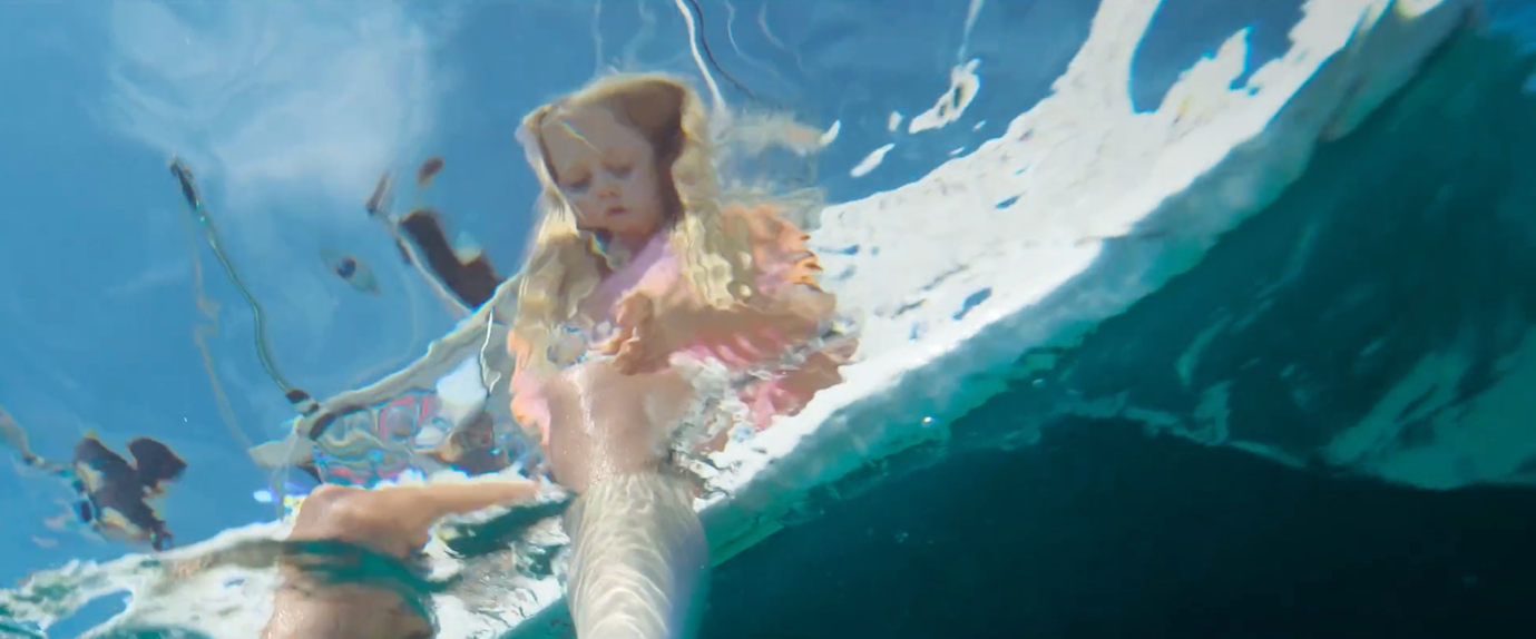 A child with long blonde hair stands partially submerged in water, their reflection distorted by ripples. The sky is visible above, along with hints of surrounding objects captured in the water's surface.