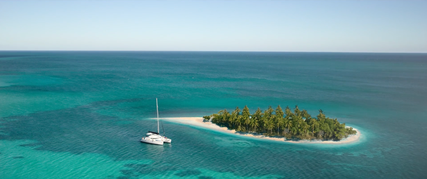 A small, lush island with palm trees is surrounded by turquoise waters, with a sailboat anchored nearby. The serene ocean stretches into the horizon under a clear sky.