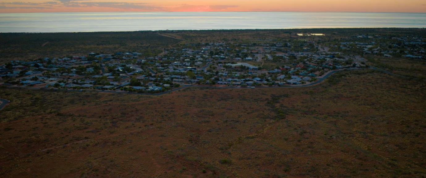 Aerial view of a small town nestled in a desert landscape, with a stretch of calm water visible along the horizon. The sky transitions from orange to purple as the sun sets, casting a warm glow over the area.