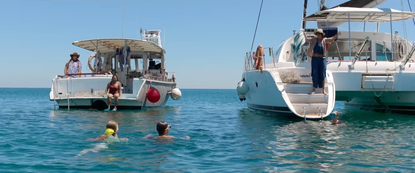 Two boats are anchored in clear, blue water under a bright sky, with several people enjoying themselves in the water nearby. One person stands on the stairs of a boat, while others swim and relax in the gentle waves.