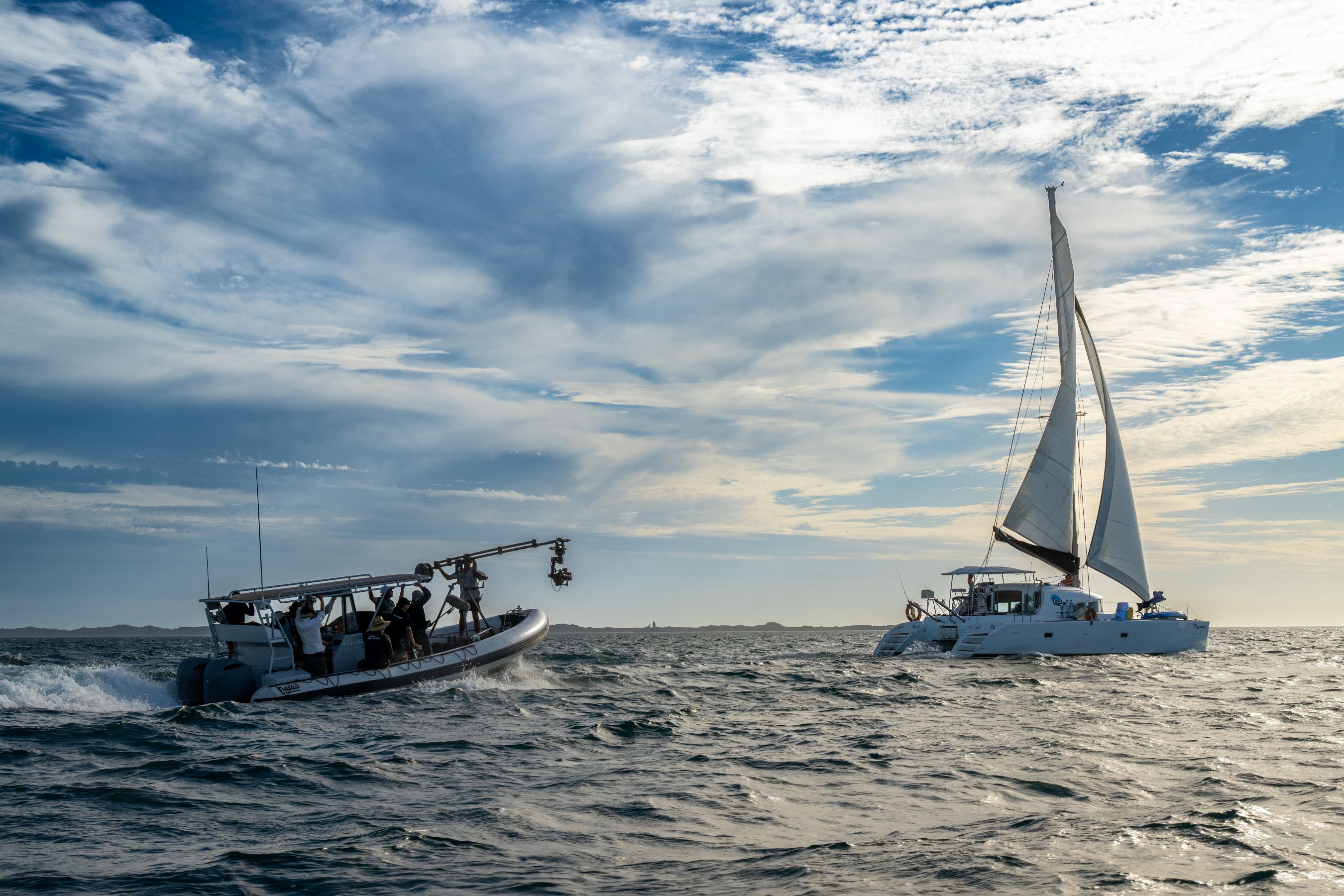 A motorboat with crew members and a mounted camera moves through ocean waves, closely trailing a larger sailing catamaran with its sails raised. The backdrop features a sky filled with clouds, casting a soft light on the serene water.
