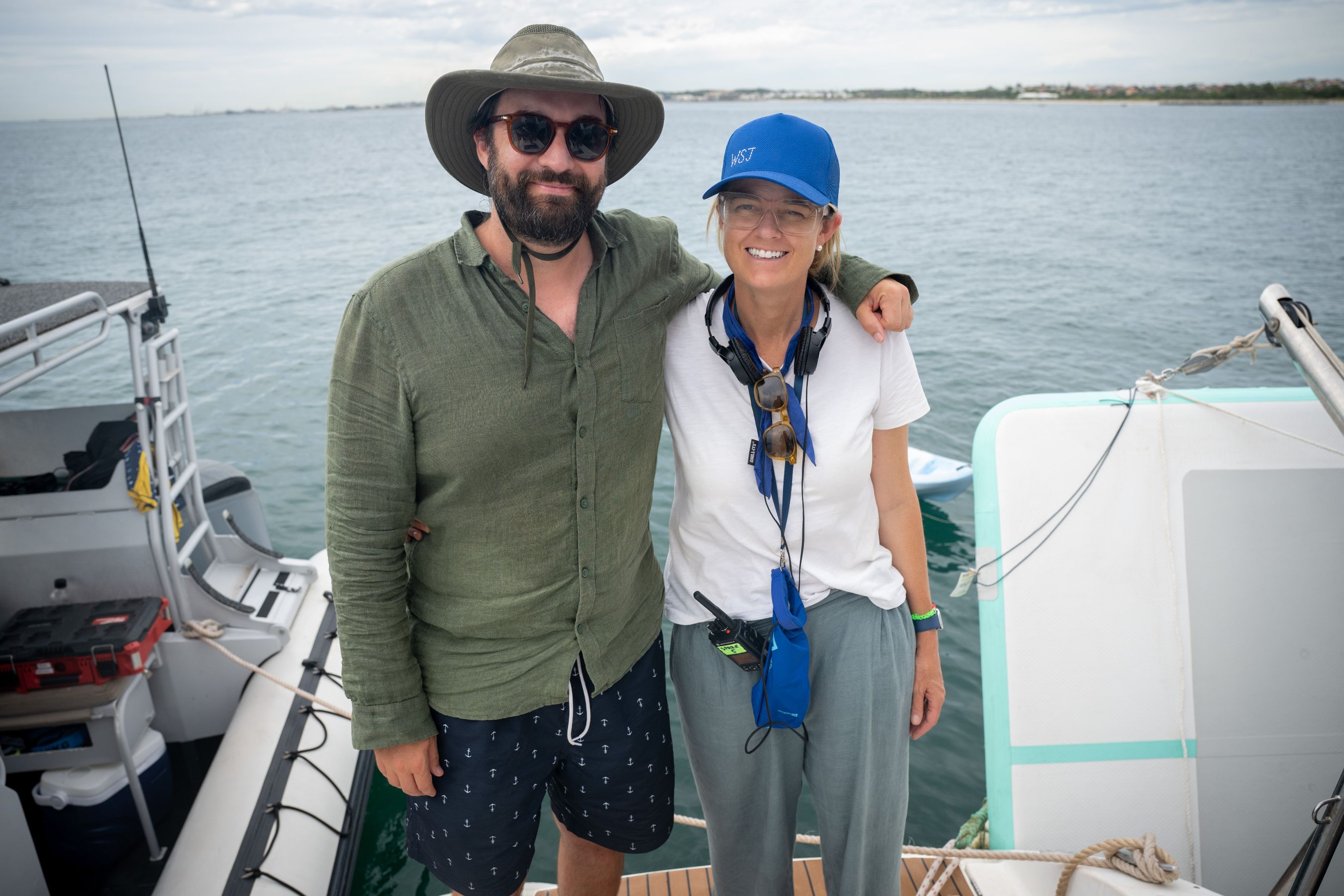 Two individuals stand together on a boat, smiling at the camera. One person wears a green shirt and sunglasses, while the other has a blue cap and a white shirt, with a headset around their neck. The backdrop features a calm body of water and a cloudy sky.