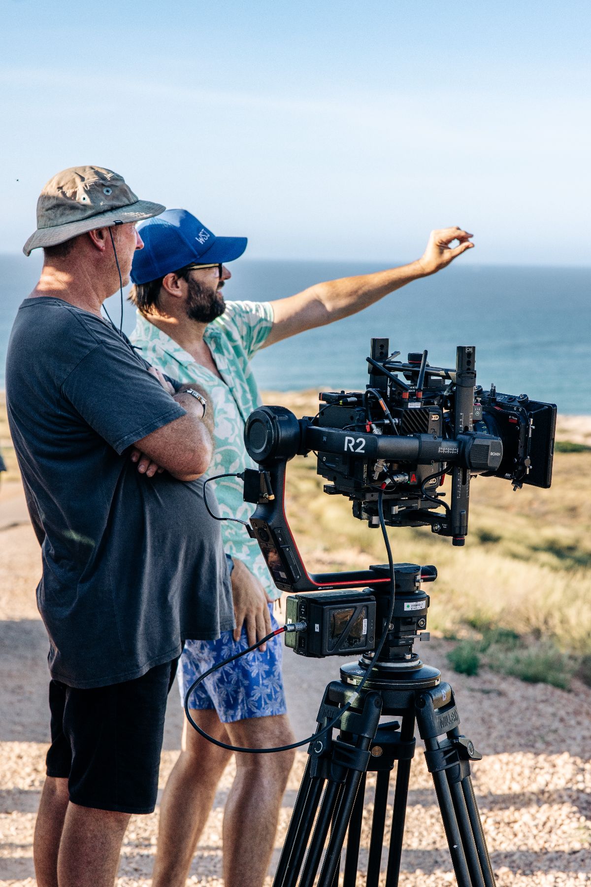 Two men stand beside a professional camera setup on a tripod, with one gesturing towards the ocean in the background. The scene is outdoors, displaying a clear sky and grassy terrain.