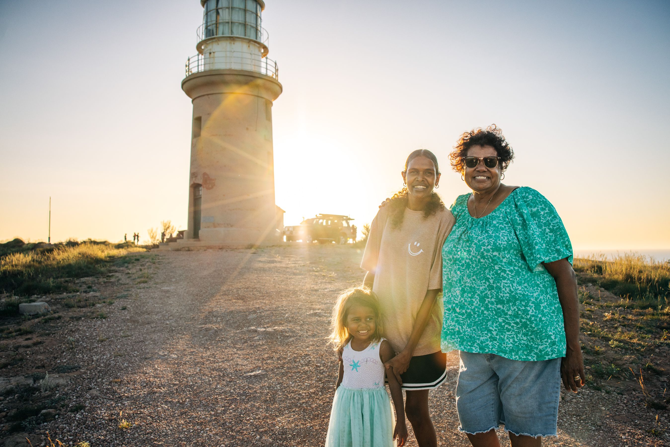 A woman and two girls stand smiling in front of a lighthouse at sunset, with warm sunlight illuminating the scene. The landscape features grassy areas and a gravel path leading to the lighthouse, creating a serene family moment.