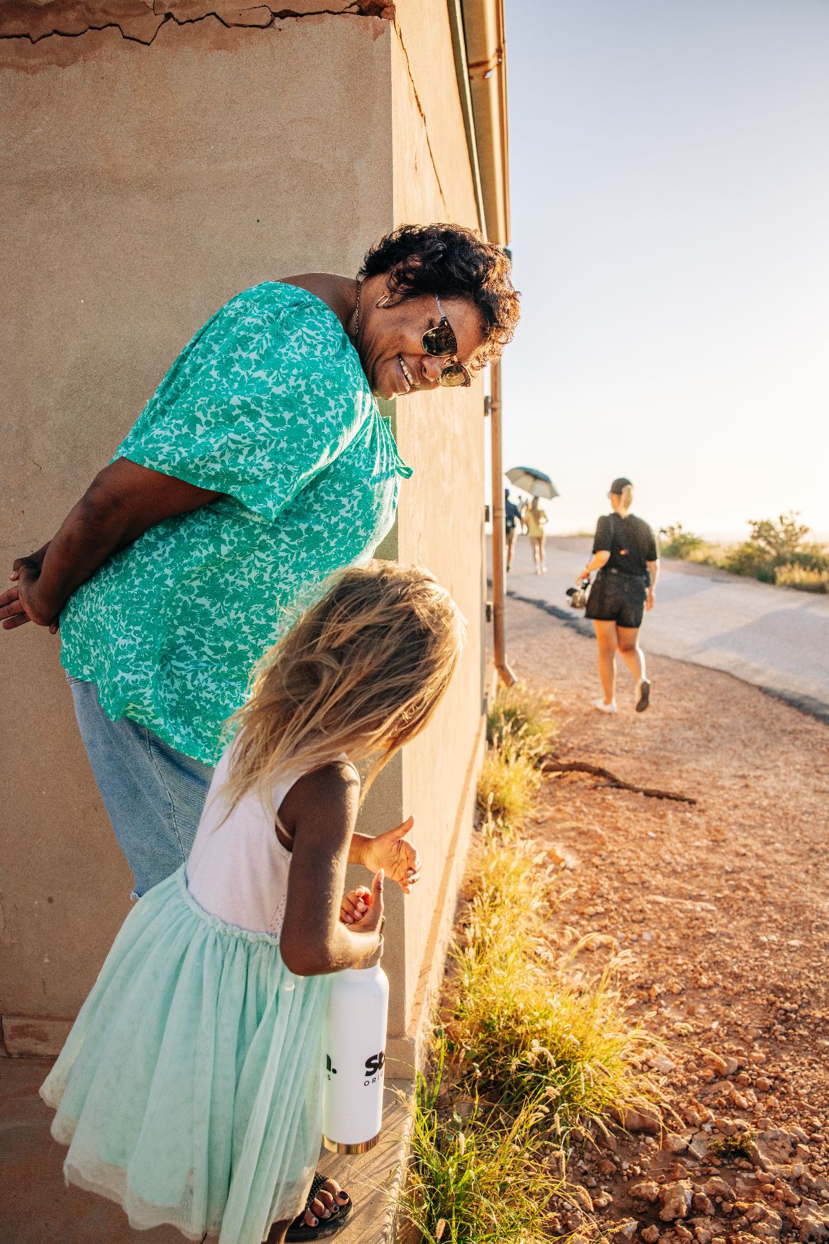 A woman in a bright green blouse smiles while leaning against a wall, interacting with a young girl wearing a light dress and holding a water bottle. In the background, other people walk along a path under warm sunlight, surrounded by grass and dirt.