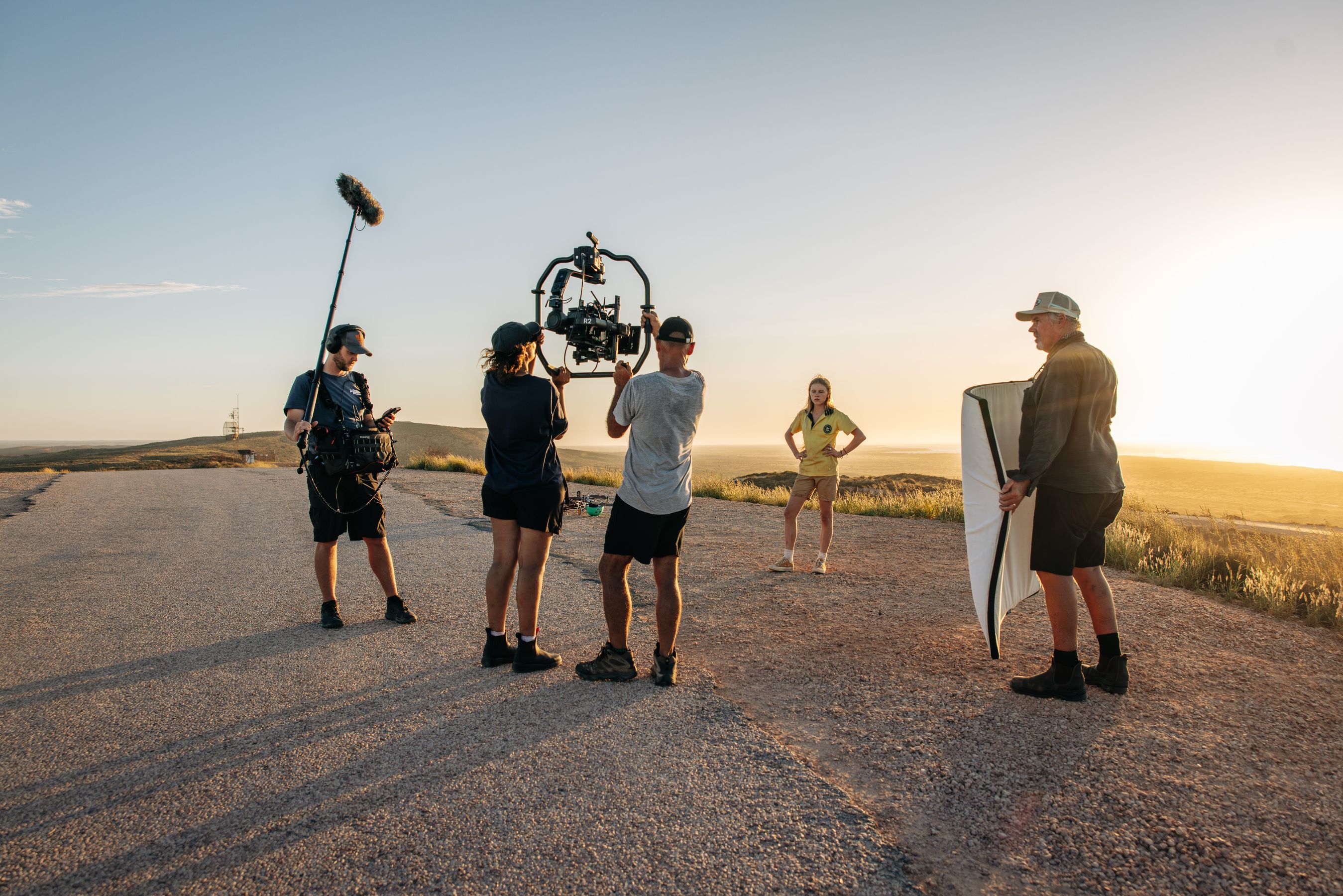 A film crew is set up on a scenic, sunlit road, with one person using a camera gimbal and another holding a microphone. In the foreground, a young woman poses confidently while another crew member adjusts a reflector to enhance the lighting. The background features rolling hills under a clear sky.