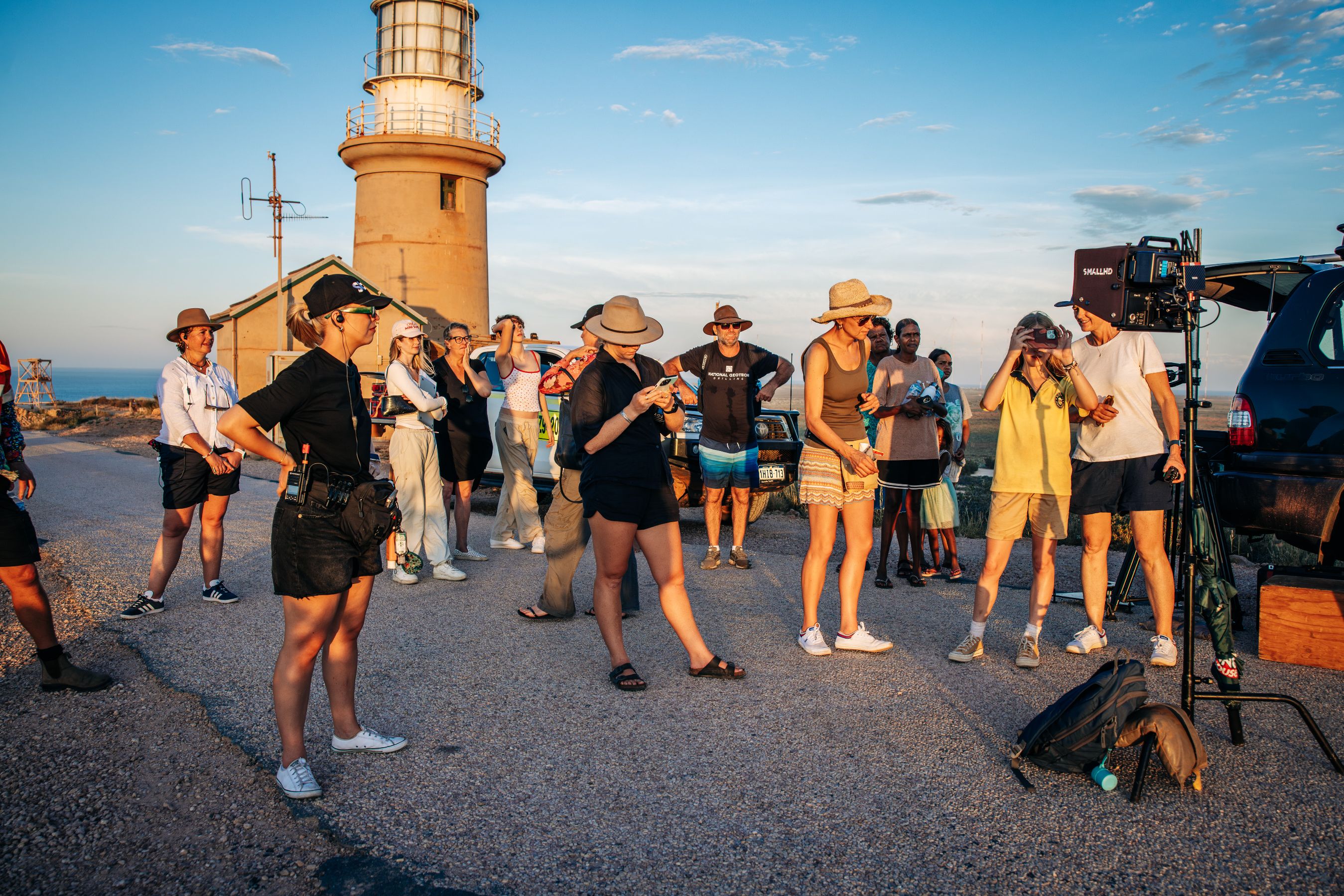 A diverse group of people gathers near a lighthouse at sunset, some taking photos while others chat and engage with one another. The scene includes various styles of clothing and accessories like hats, and a vehicle is parked nearby, contributing to a relaxed atmosphere in a coastal setting.