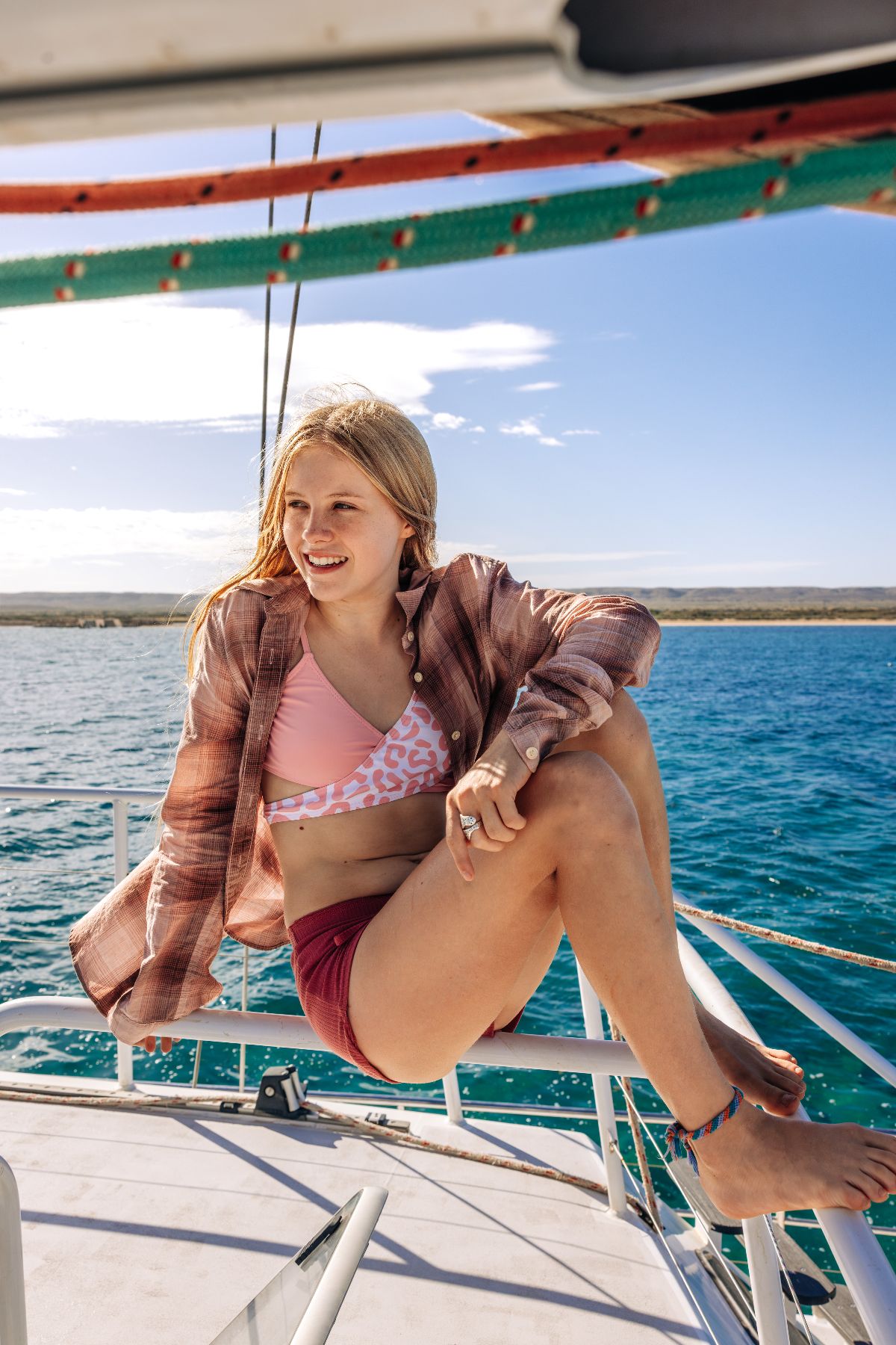 A young woman sits on the edge of a boat, smiling and looking out over the water. She wears a pink bikini top with a leopard print, paired with reddish shorts, and a loosely buttoned plaid shirt. The background features a clear blue sky and calm sea.