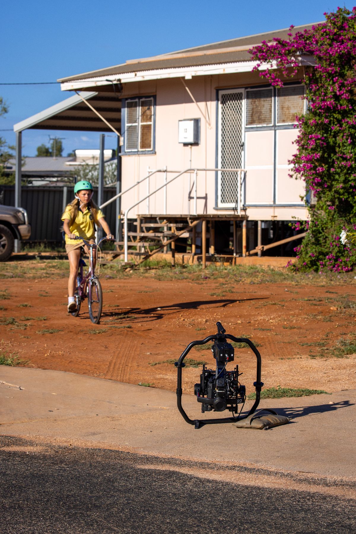A girl in a yellow shirt and green helmet rides a bicycle along a dirt path in front of a house with a porch and blooming bougainvillea. A camera on a stabilizer stands on the ground, positioned to capture her ride.
