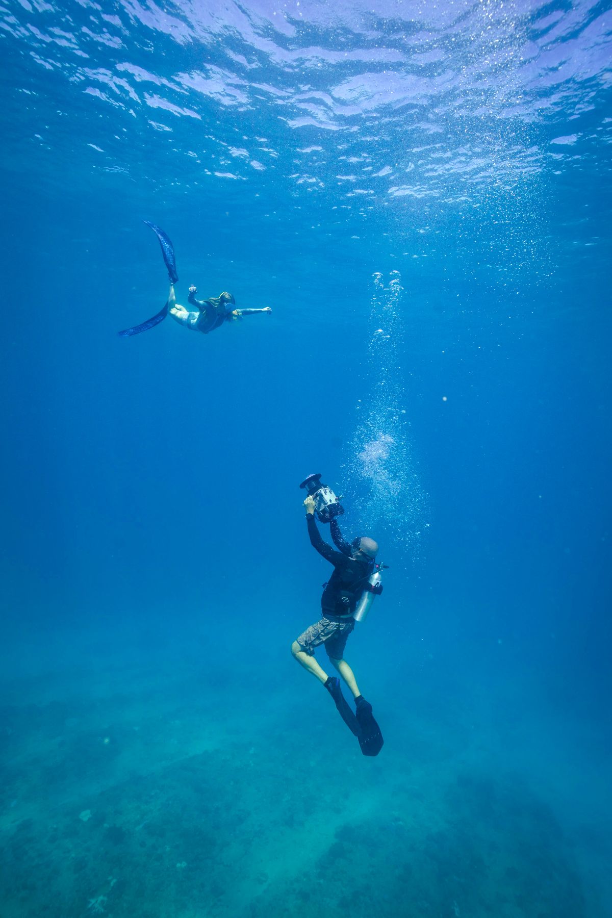 Two divers are underwater in a clear blue ocean; one is swimming with fins, while the other is taking photos with an underwater camera. Bubbles are rising around them, and the ocean floor is visible below.