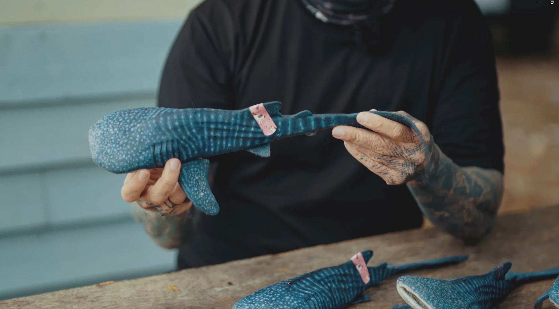 A person with tattooed hands holds a small, blue, plush whale shark, inspecting it closely. Several similar plush whale sharks are arranged on a wooden table in front of them.