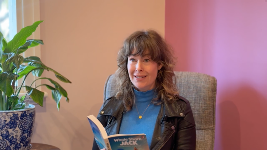 A woman with wavy brown hair sits in a cozy chair, smiling while holding a book titled "Whale Shark Jack." She wears a leather jacket over a blue turtleneck, and a green plant is in a decorative pot nearby, with a softly colored wall in the background.