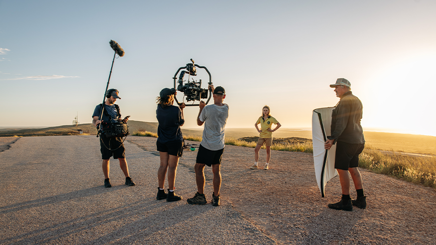 A film crew is set up on a rural road during golden hour, with a female model posing for the camera. One crew member holds a boom microphone, while others operate cameras and adjust lighting reflectors. The landscape features rolling hills and a clear sky, creating a warm, radiant ambiance.