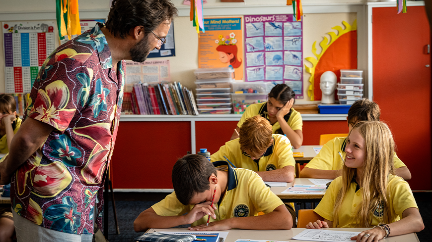 A teacher in a colorful floral shirt interacts with a classroom of students wearing yellow shirts. Some students are laughing or covering their faces while others are engaged in their work, creating a lively and humorous atmosphere.