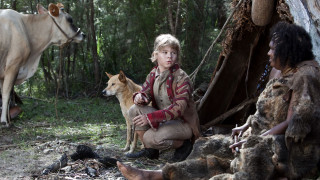 A young child sits on the ground beside a dingo, looking intently towards a nearby cow. An adult person is seated nearby, partially covered in fur, with a fire pit visible between them in a forested setting.