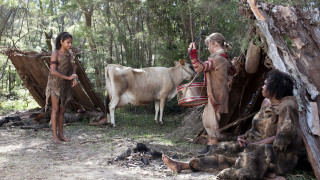 A young boy in primitive attire stands near a cow, while a girl in similar clothing plays a drum and appears to be engaging with the cow. Two other individuals, one sitting and another partially hidden, are near makeshift shelters made of branches and leaves in a forested area.