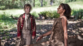 A young boy in a red and beige military-style jacket stands in a forest clearing, looking at a girl dressed in a tattered, brown garment. The girl, with her hair down and a joyful expression, stands beside a fallen log, surrounded by greenery and sunlight filtering through the trees.