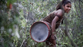 A young girl stands in a dense, green forest, wearing a simple, earth-toned outfit. She is smiling and holding a large drum, positioning it as if ready to play. The surrounding vegetation includes tall grasses and small shrubs, creating a lively natural backdrop.