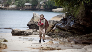 A young boy in a brown shirt and shorts runs along a sandy beach, surrounded by rocky formations and shallow water. Lush greenery is visible in the background, and the scene conveys a sense of adventure and playfulness.